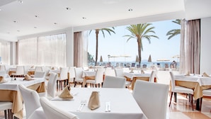Un restaurante con mesas dispuestas para comer, ventanas amplias con cortinas y vistas a la playa.