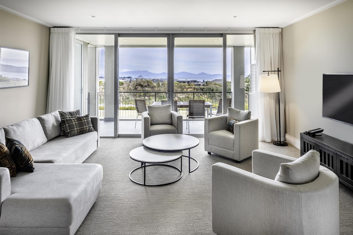 Un salon moderne comprenant un canapé, des fauteuils, une table basse et un téléviseur. Il est doté de grandes fenêtres offrant une vue sur les montagnes et d’un balcon.