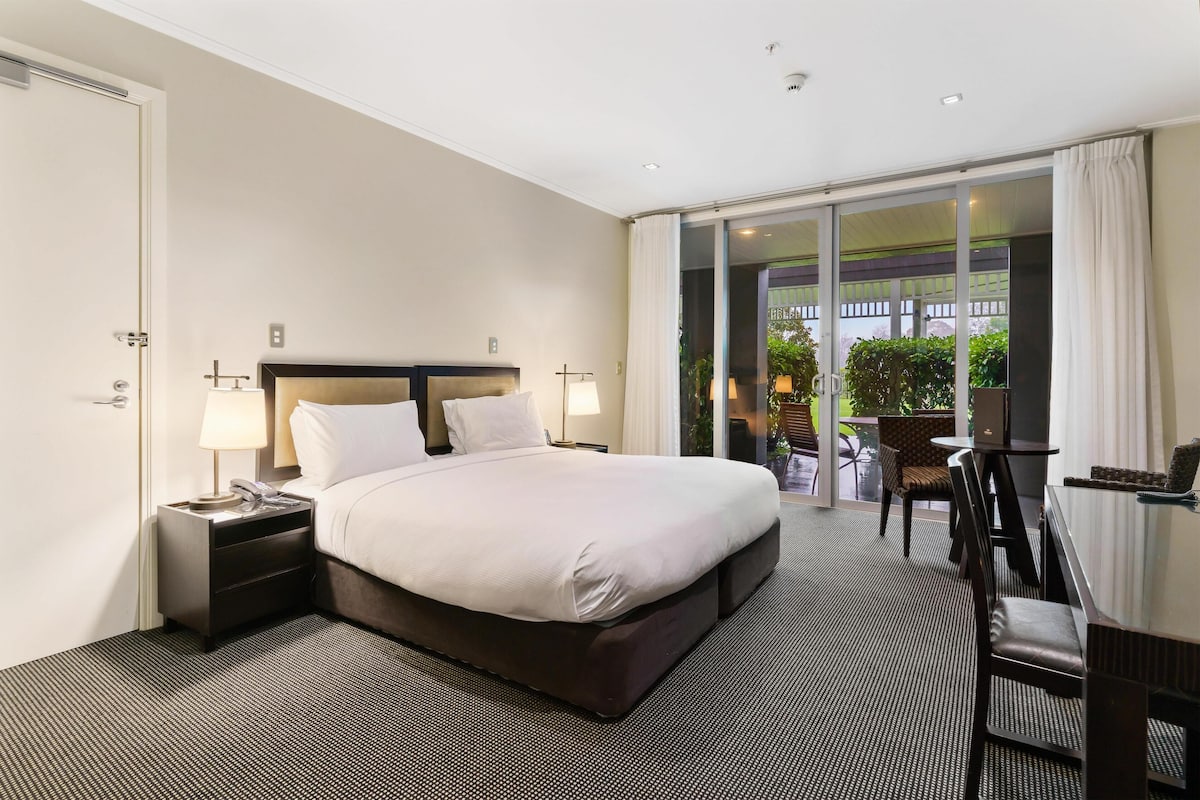 Une chambre d’hôtel avec un grand lit, un bureau et une chaise. Une porte-fenêtre coulissante donne accès à un espace extérieur agrémenté de plantes.