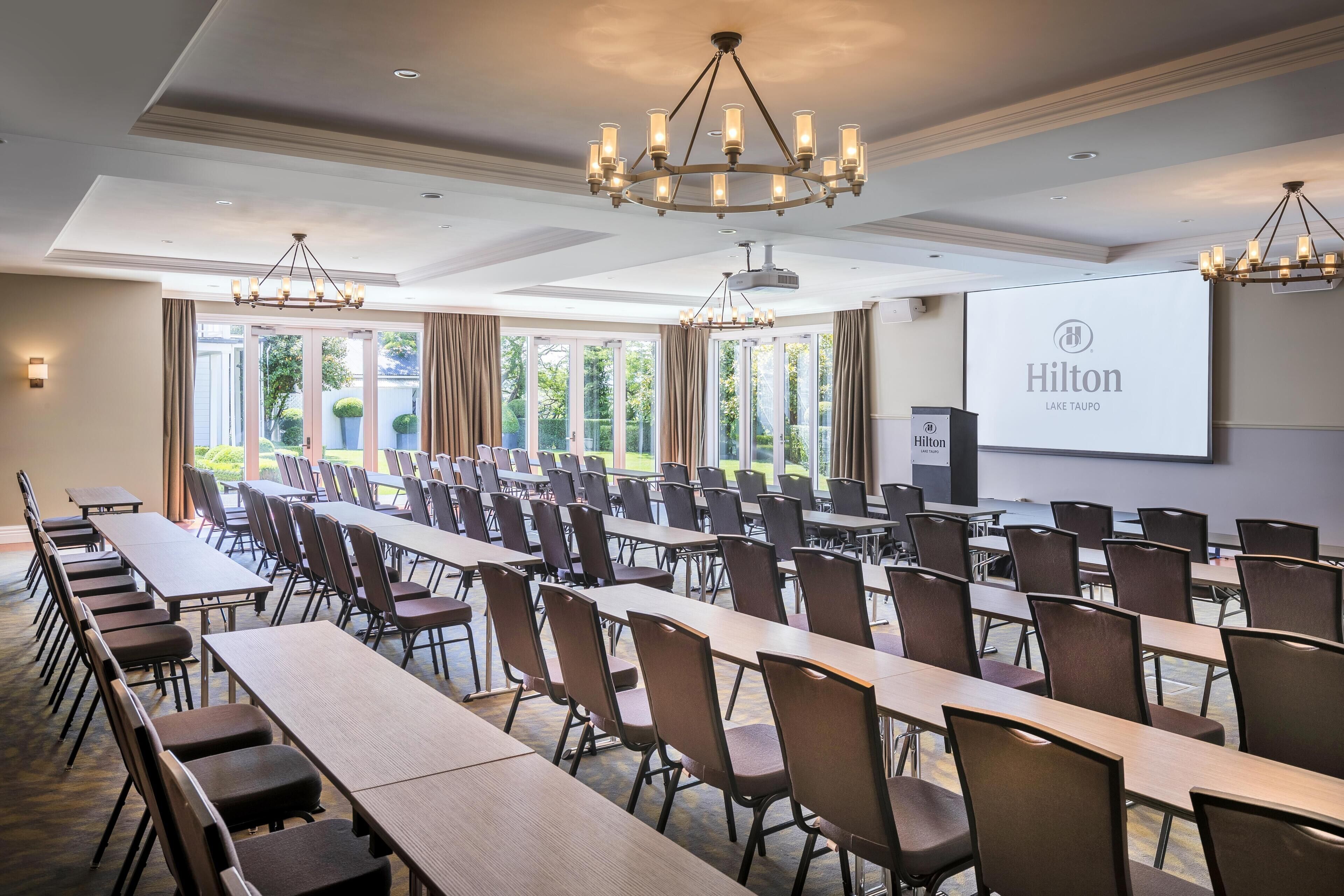 Ein Konferenzraum mit langen Tischen und Stühlen, großen Fenstern und einer Projektionsleinwand mit dem Hilton-Logo.