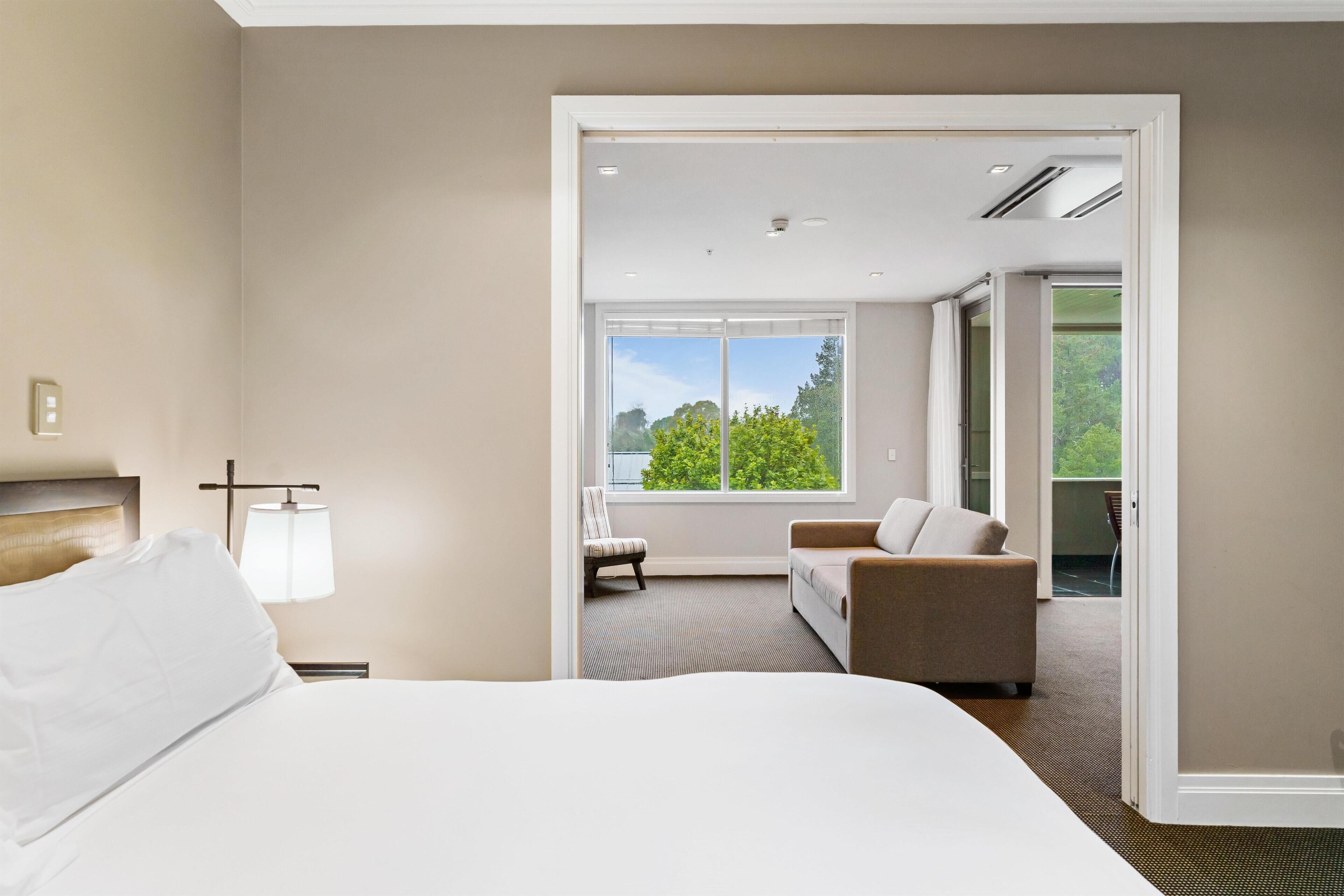 Une chambre d’hôtel moderne dotée d’un grand lit, d’un coin salon avec un canapé et un fauteuil, d’une fenêtre donnant sur un espace verdoyant et d’une porte menant à une autre pièce.