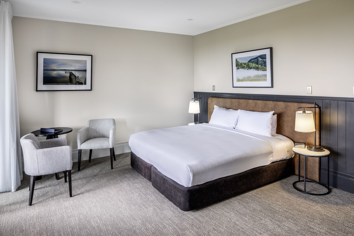 Une chambre d’hôtel avec un grand lit, deux fauteuils, une petite table, deux lampes fixées au mur et deux tableaux encadrés.