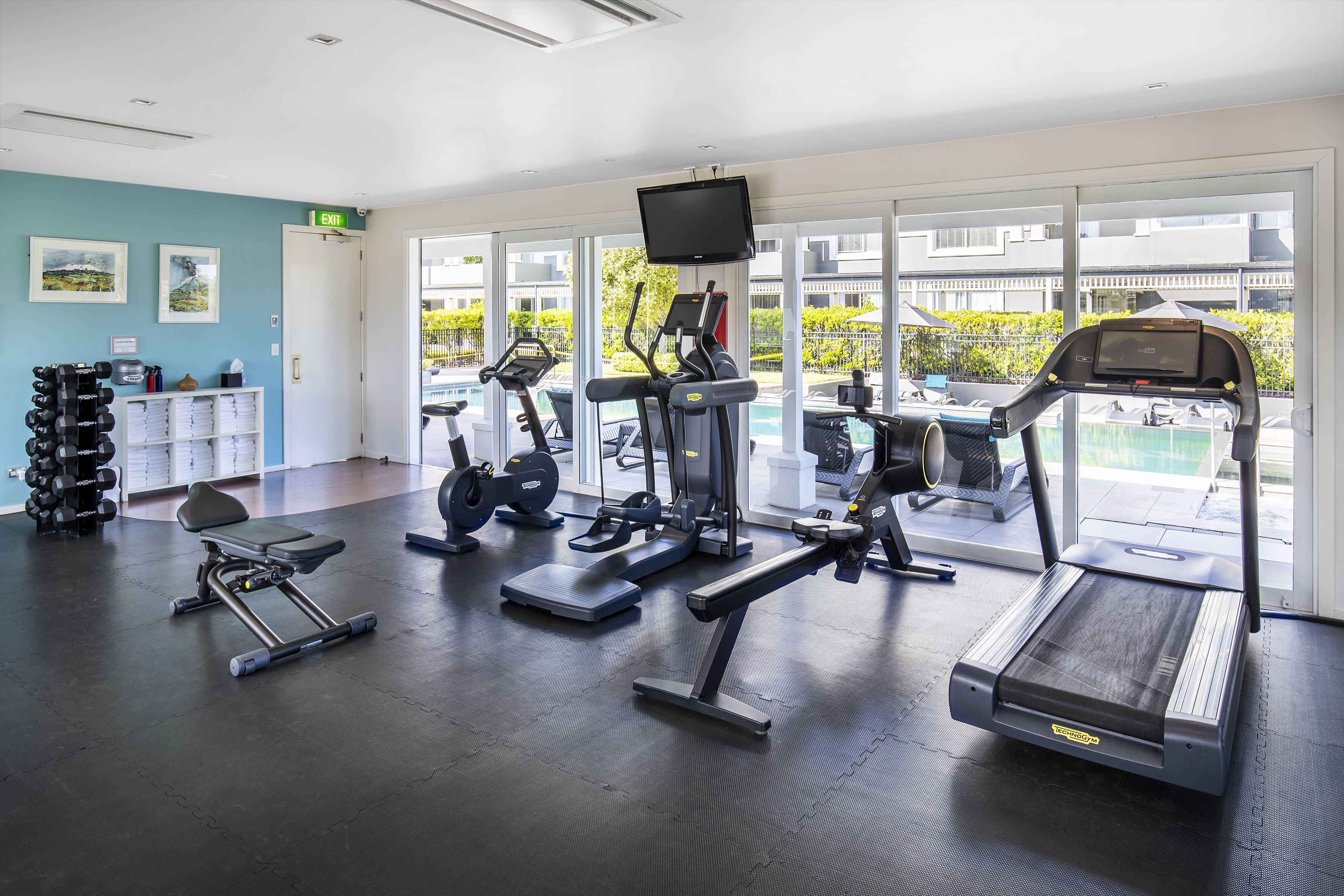 Ein Fitnessstudio mit Laufbändern, Kraftgeräten und Kurzhanteln, große Fenster mit Blick auf einen Pool und Gebäude.