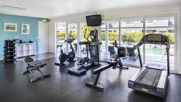 Un gimnasio con cintas de correr, máquinas de pesas y mancuernas, ventanas amplias con vistas a una piscina y edificios.