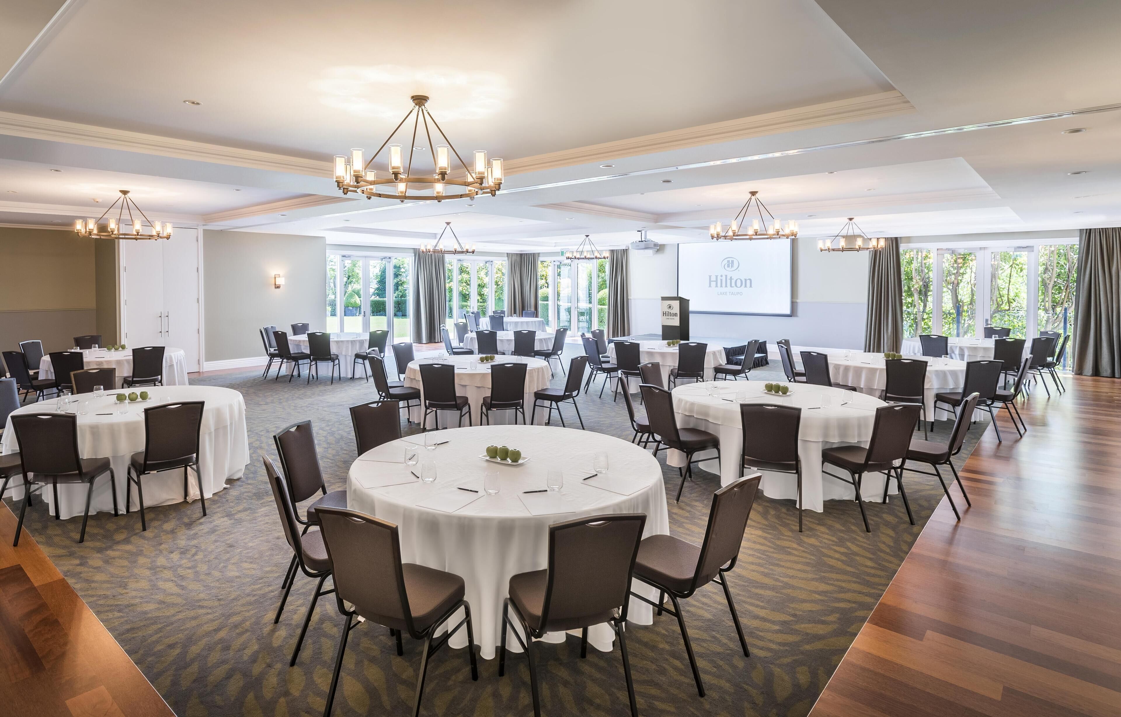 A large, well-lit conference room with multiple round tables, each set with chairs and white tablecloths. There are chandeliers hanging from the ceiling, and a projector screen is visible on the wall.