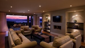 A modern living room with a large sofa set, a coffee table, and a flat-screen TV mounted on the wall. There is a fireplace with decorative items and a view of the outdoors through large windows.