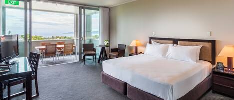 A hotel room with a large bed, a desk with a chair, a small table with a chair, and a balcony with a view.