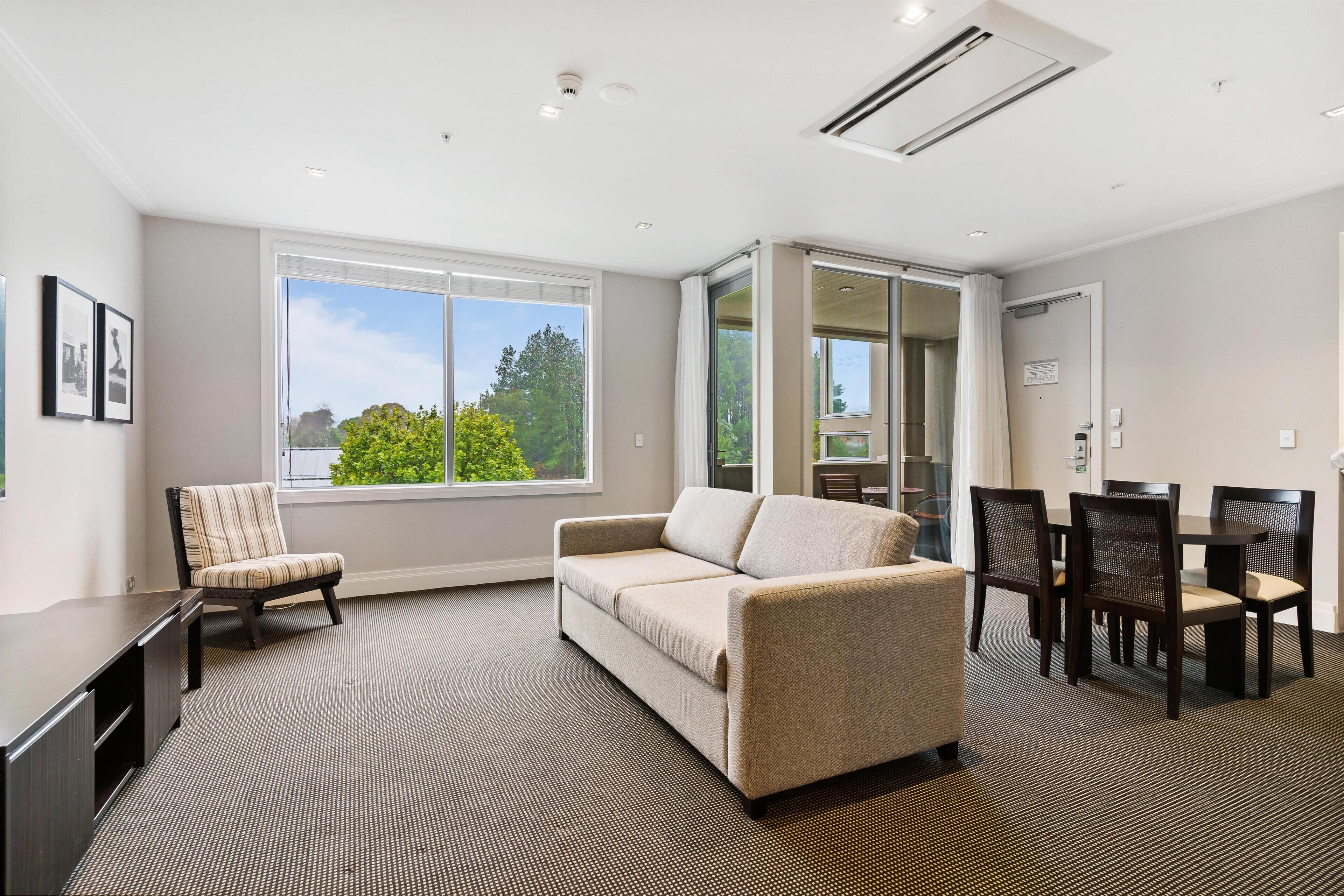 A modern hotel room with a sofa, a chair, a dining table with chairs, a desk, and a large window offering a view of trees.