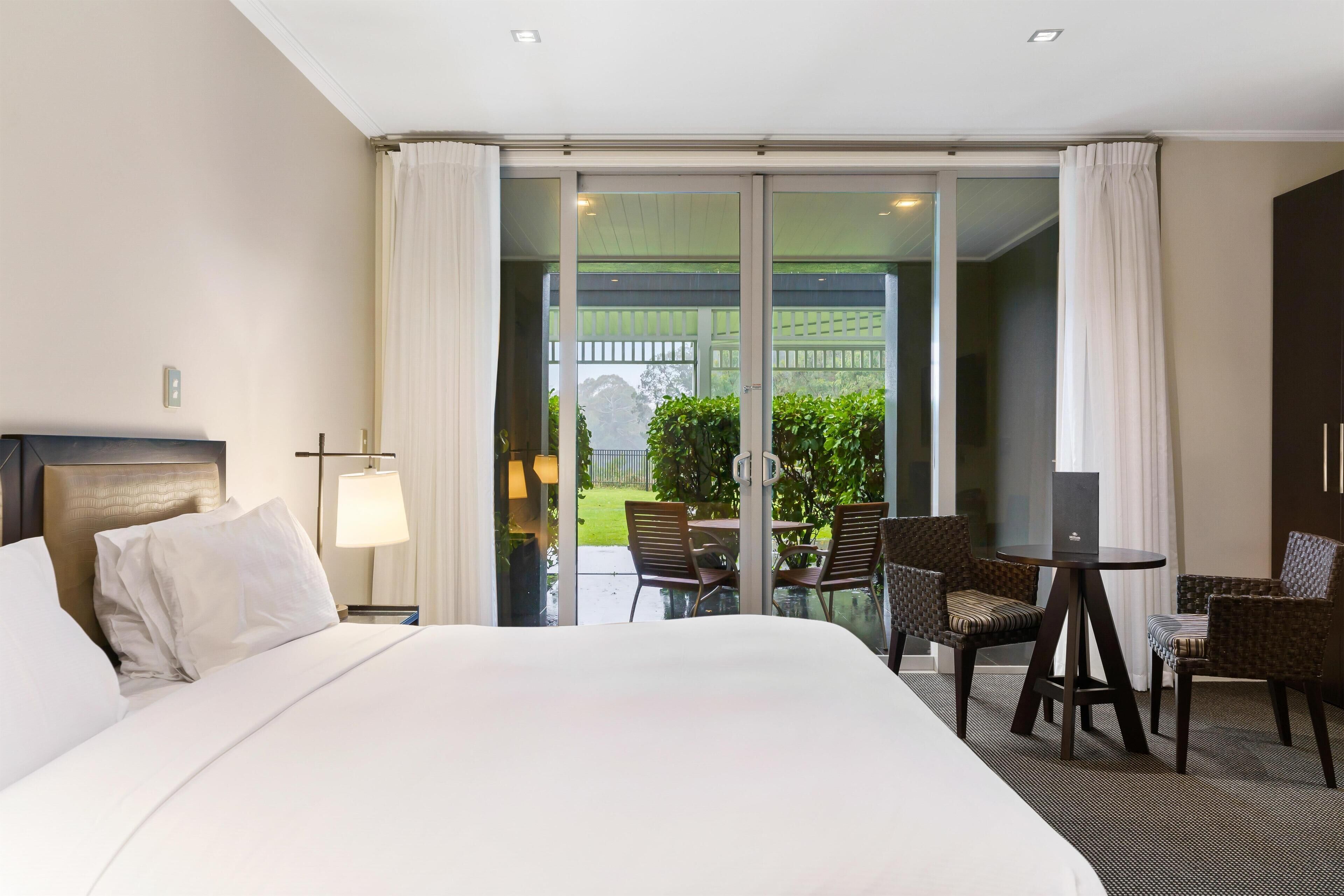 A hotel room with a large bed, a desk with two chairs, a laptop, and a view of a garden through a sliding glass door.