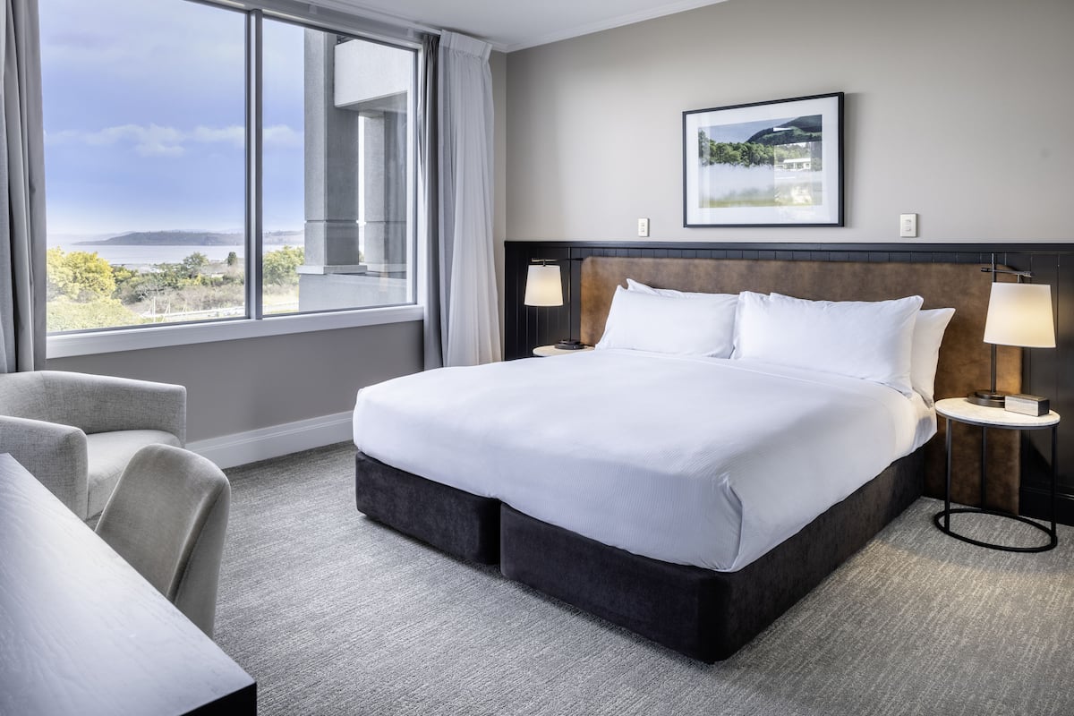 Une chambre d’hôtel avec un grand lit, deux tables de chevet avec des lampes, une chaise, une vue sur l’extérieur et un tableau encadré au mur.