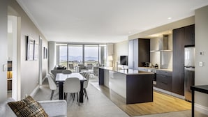 A modern hotel room with a dining area, kitchen, and a large window offering a city view.