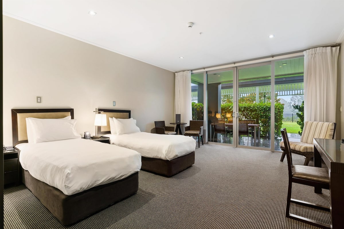 Une chambre d’hôtel avec deux lits, un bureau et une chaise. Elle dispose d’un balcon avec vue sur la verdure.