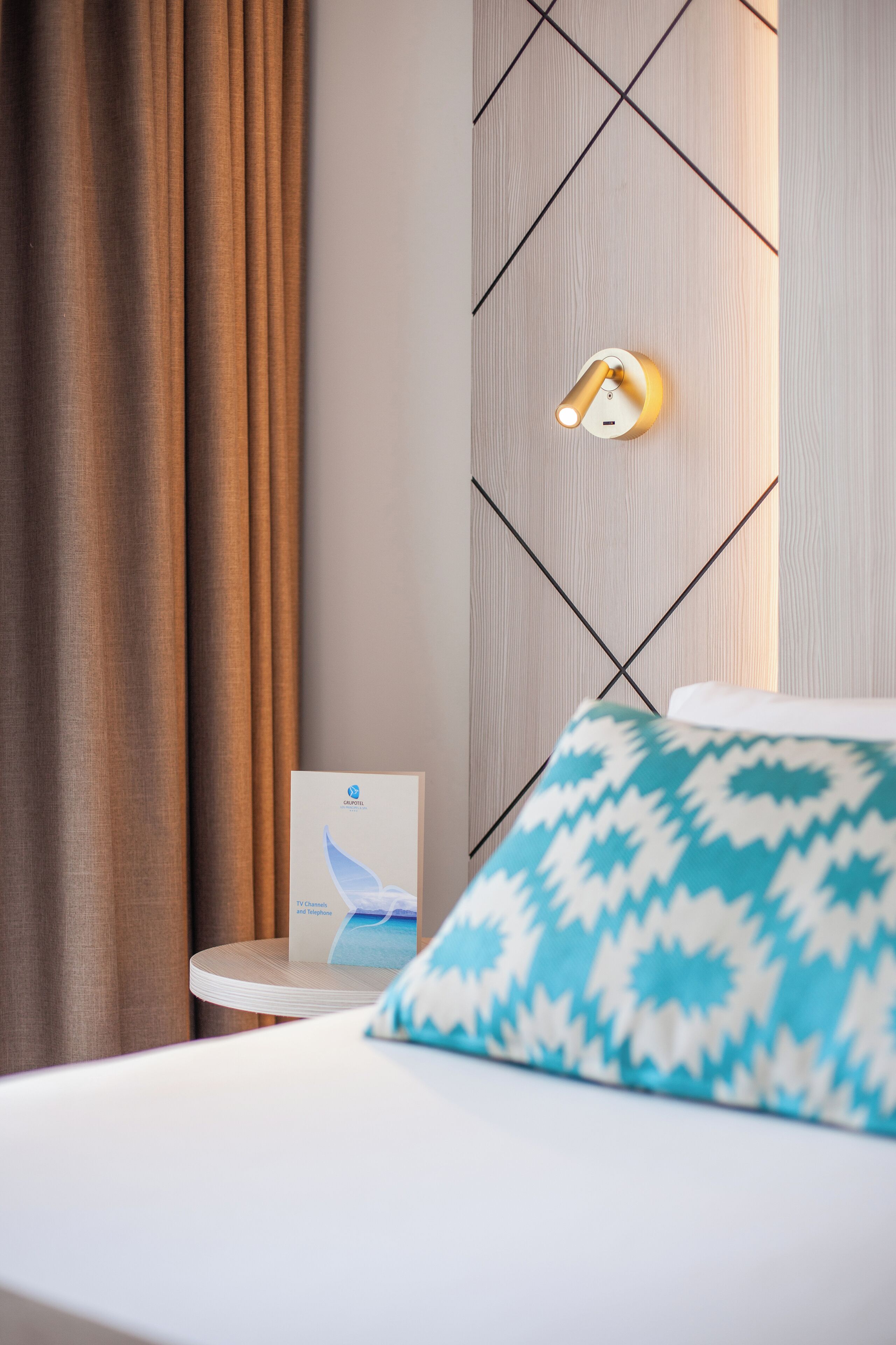 A hotel room with a bed, a wall-mounted light fixture, a small table with a brochure, and a curtain.