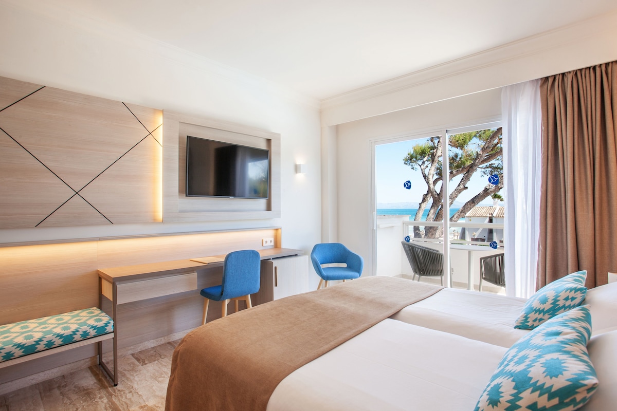 A modern hotel room with a bed, desk, and chairs, a TV mounted on the wall, and a balcony with a view of the sea and trees.