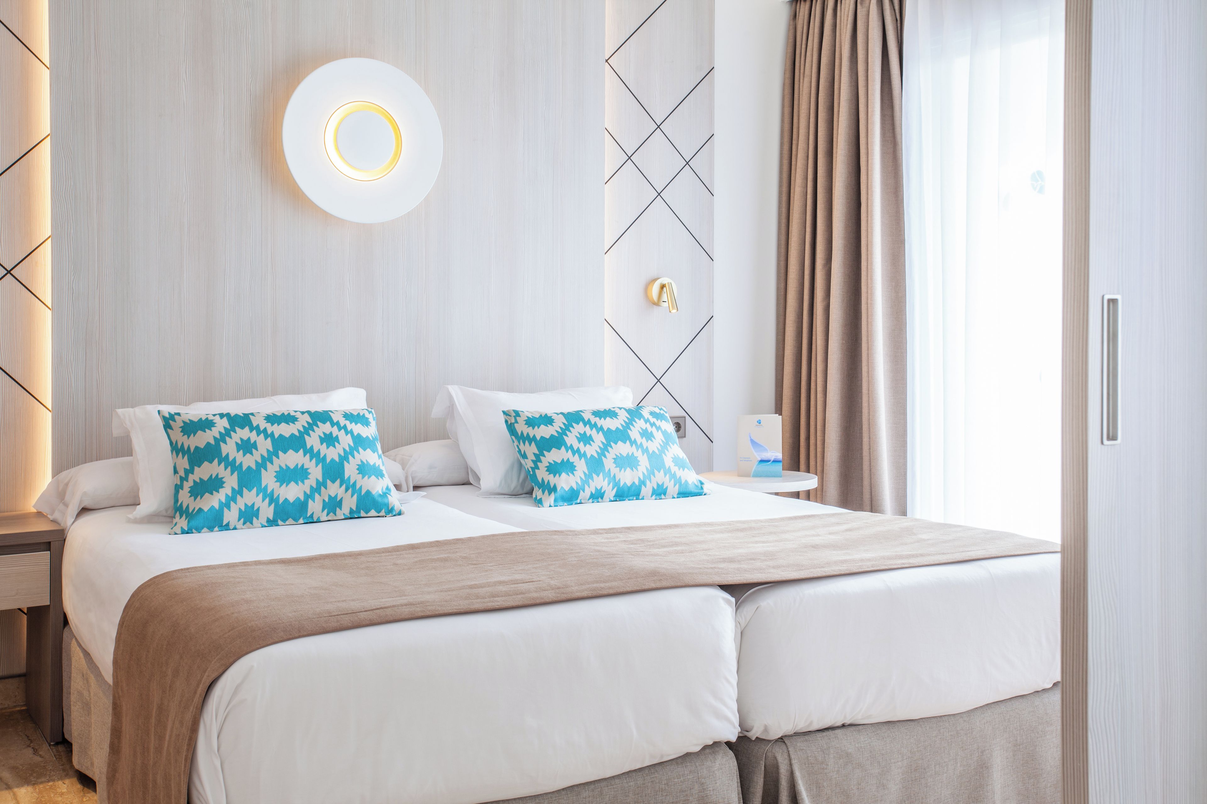 A modern hotel room with a large bed, two pillows, a bedside table, a wall-mounted light, and a window with curtains.