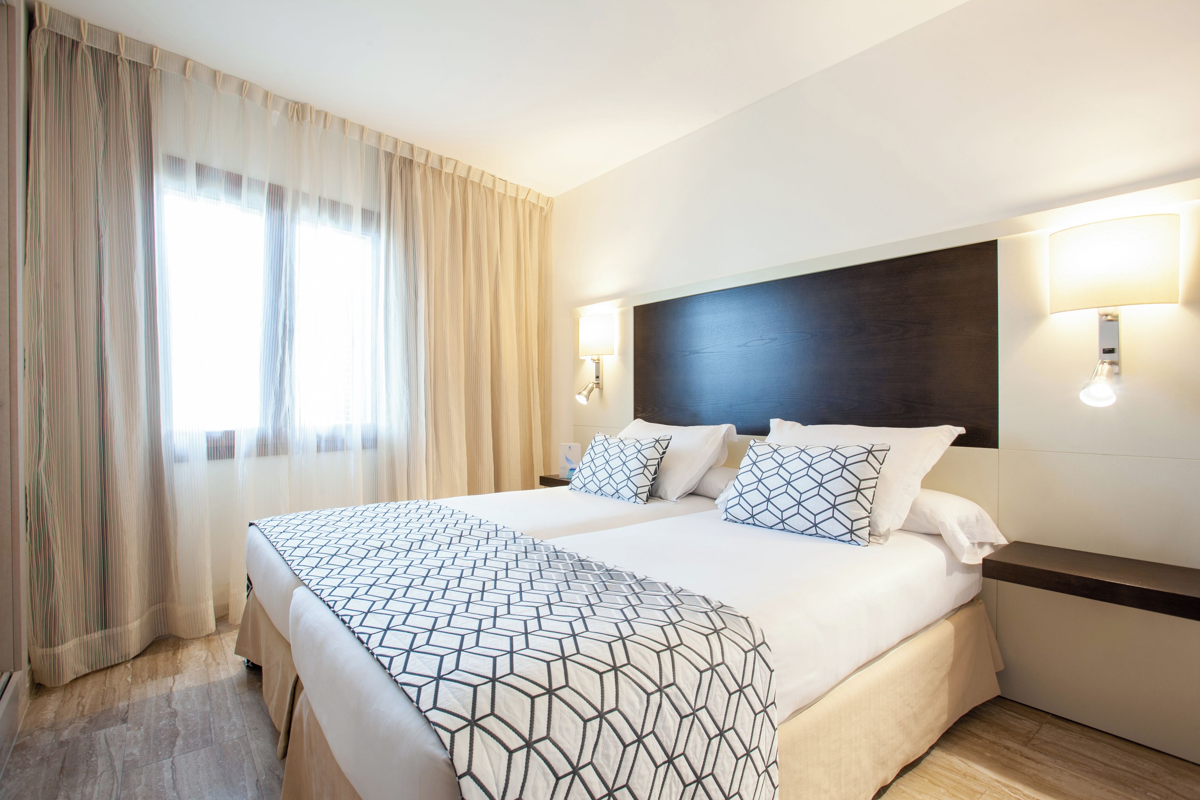 A hotel room with a large bed, two bedside tables, a headboard, and a window with curtains.