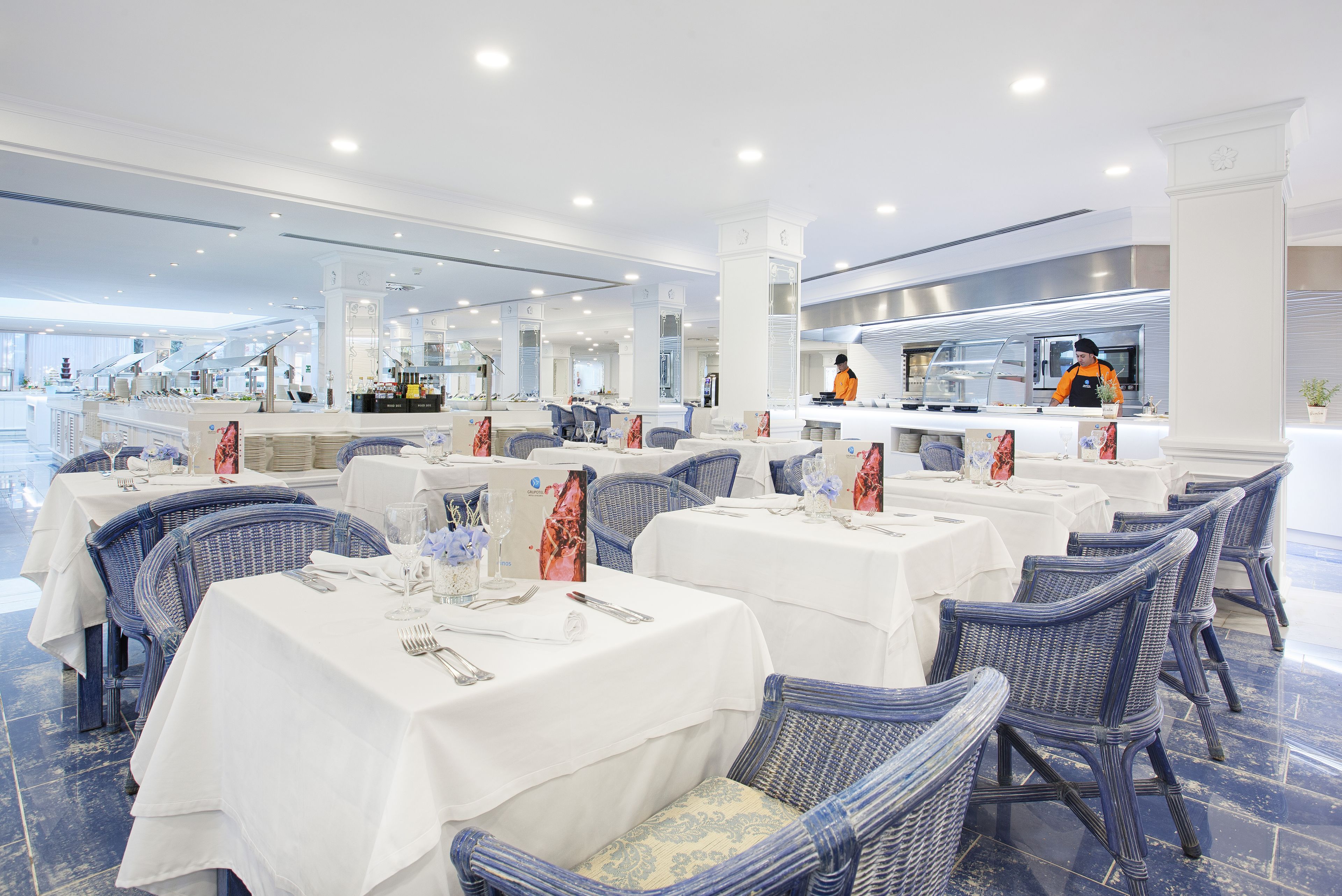 Un restaurant avec des nappes blanches, des chaises en osier et une cuisine moderne en arrière-plan.