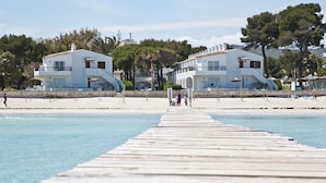 Un muelle de madera que lleva a una playa con edificios blancos y árboles al fondo.