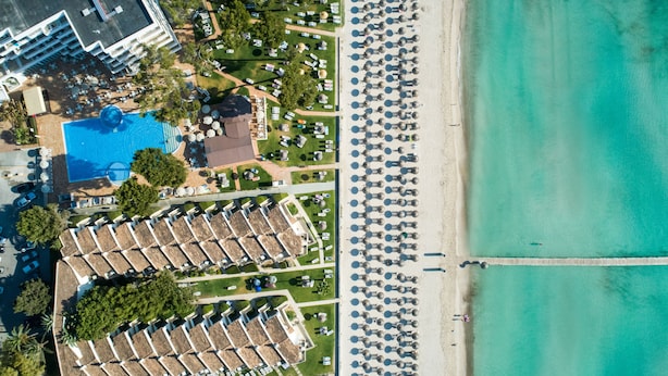 Vista aérea de un complejo turístico con una gran piscina, múltiples sombrillas de playa y un muelle largo que se extiende al agua.