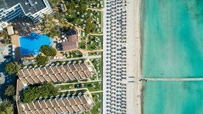 An aerial view of a resort with a large pool, multiple beach umbrellas, and a long pier extending into the water. - Grupotel Los Príncipes & Spa (Muro)