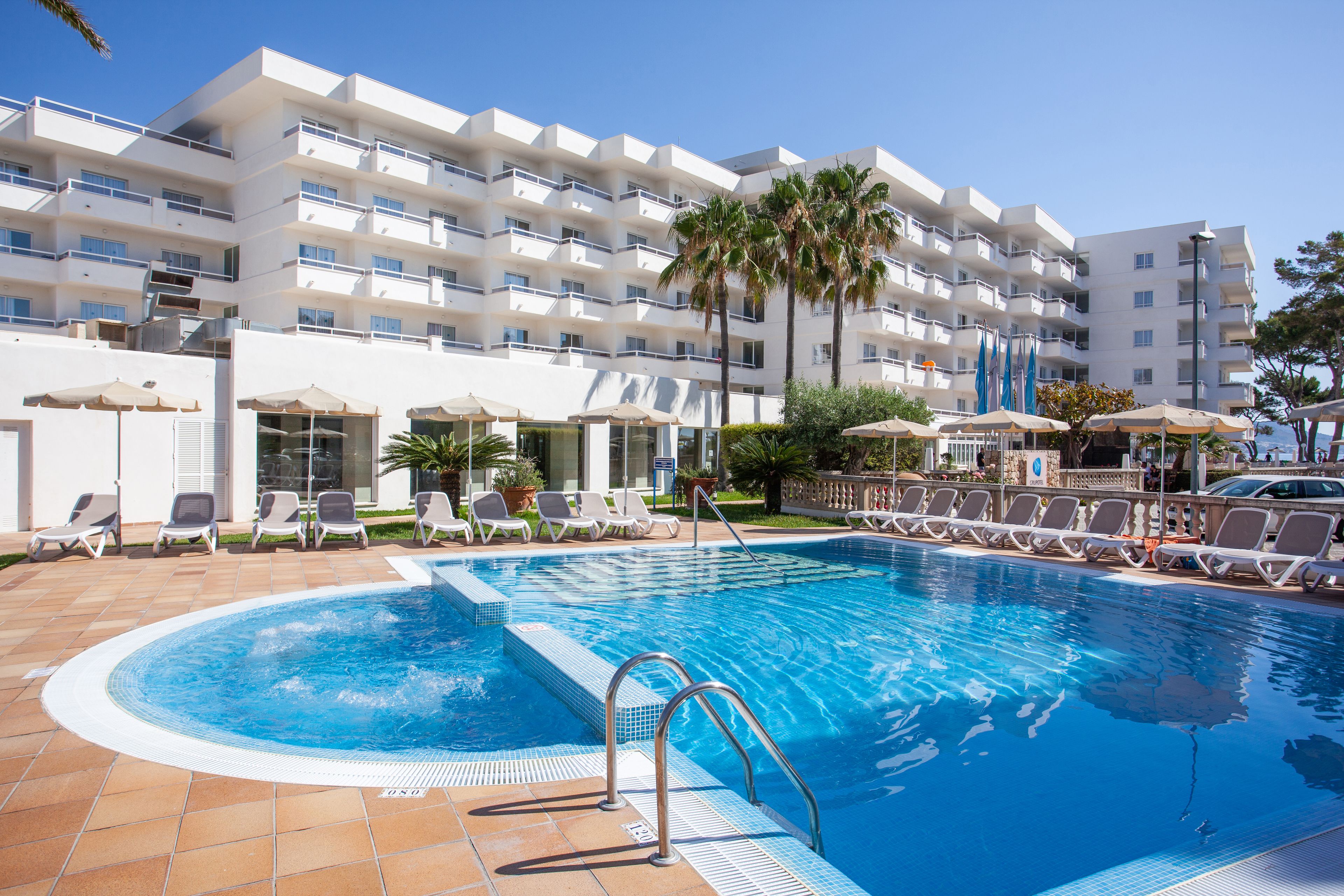 A swimming pool with lounge chairs and umbrellas, surrounded by a multi-story building with balconies.