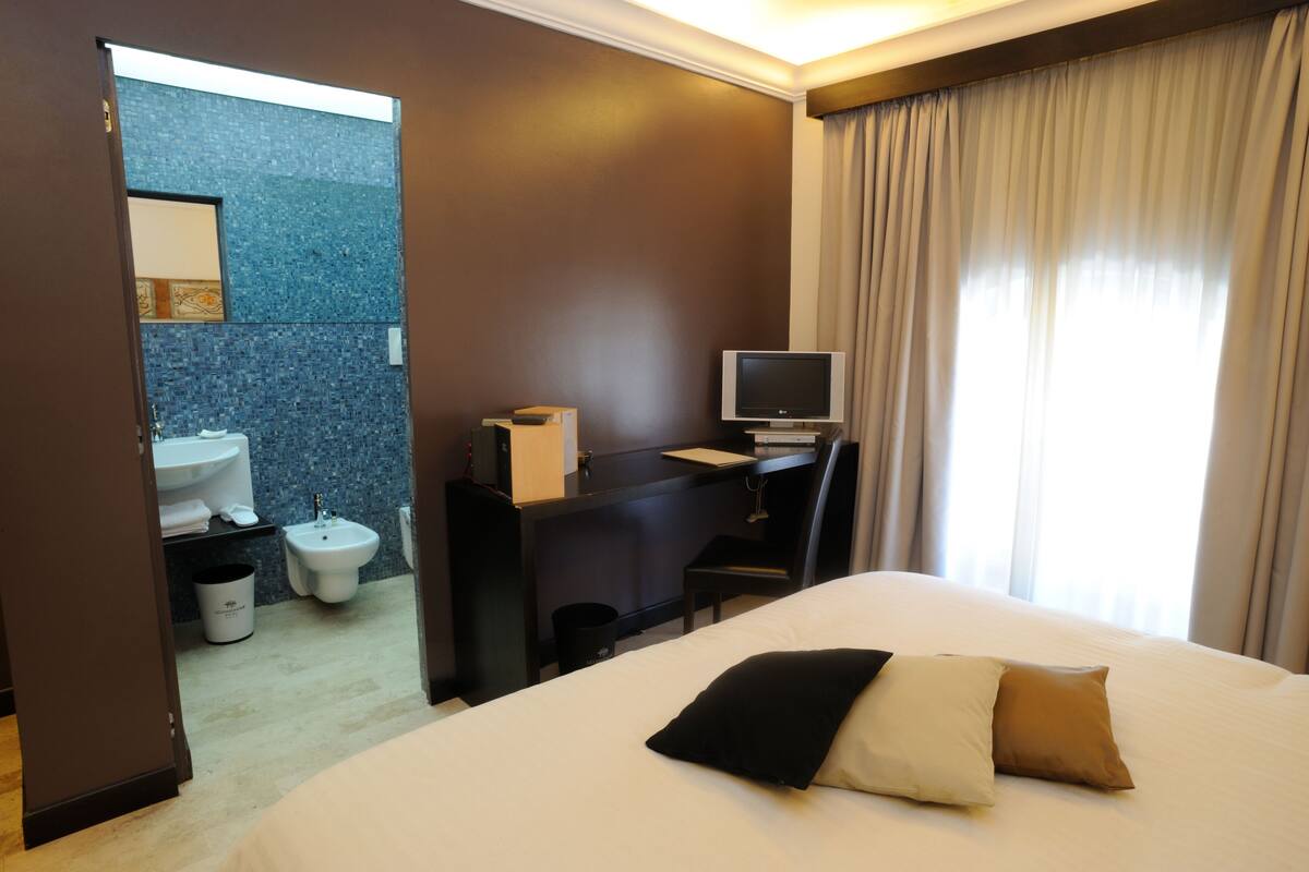Habitación de hotel con cama, escritorio con computadora, televisión, baño con lavamanos e inodoro, y ventana con cortinas.