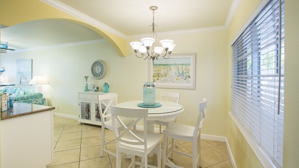 A dining area with a round table, white chairs, a chandelier, and a window with blinds.