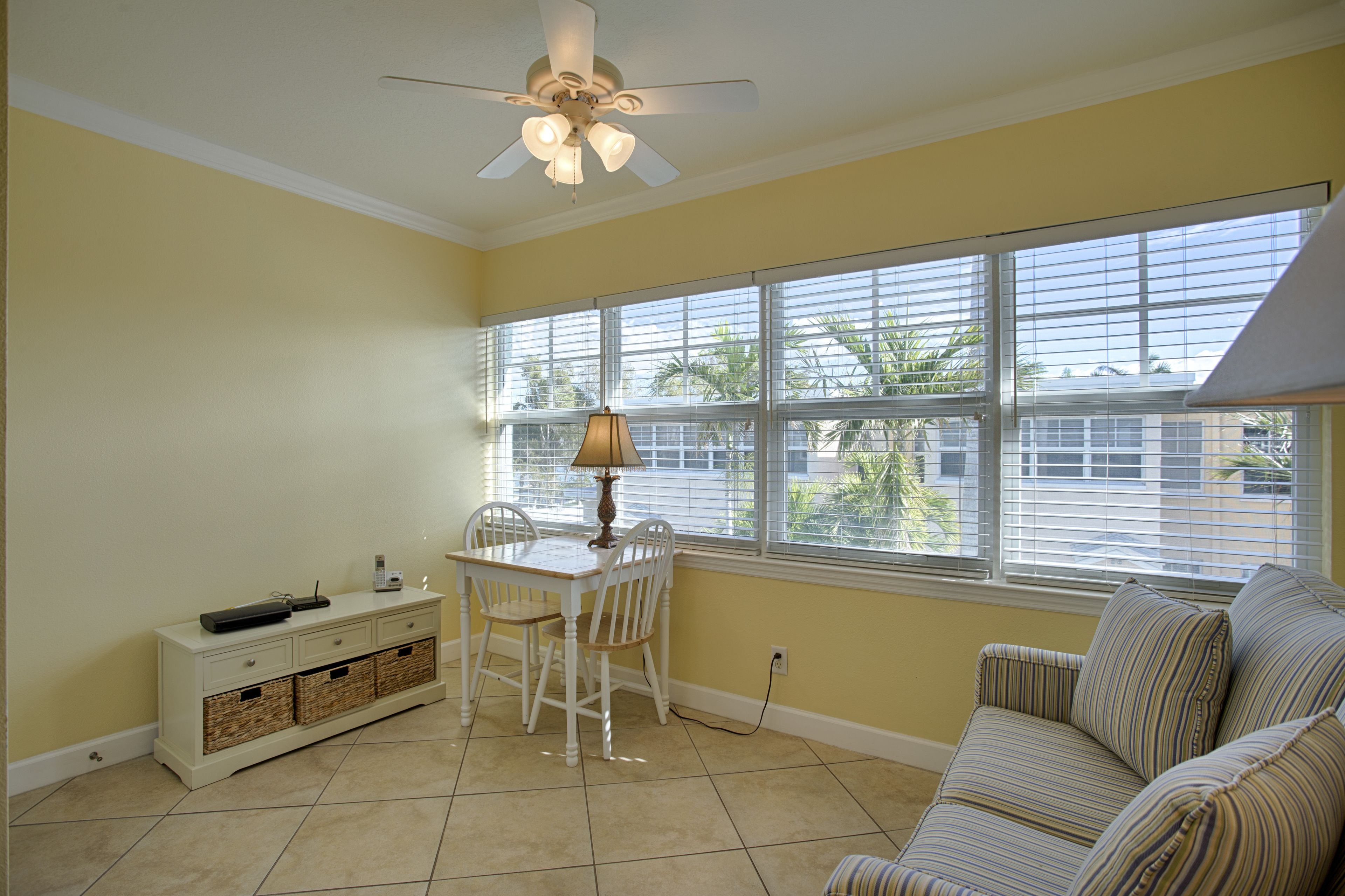 A room with a dining table, chairs, a sofa, a ceiling fan, and large windows with blinds.