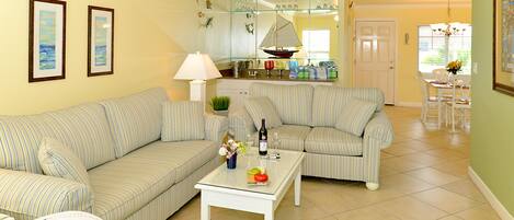 A living room with two striped sofas, a coffee table with a bottle and glasses, and a dining area with a table and chairs.
