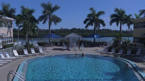 A swimming pool with lounge chairs and palm trees, surrounded by buildings and a view of a lake.