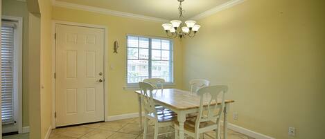 A dining area with a wooden table and white chairs, a chandelier, a door, and a window with blinds.