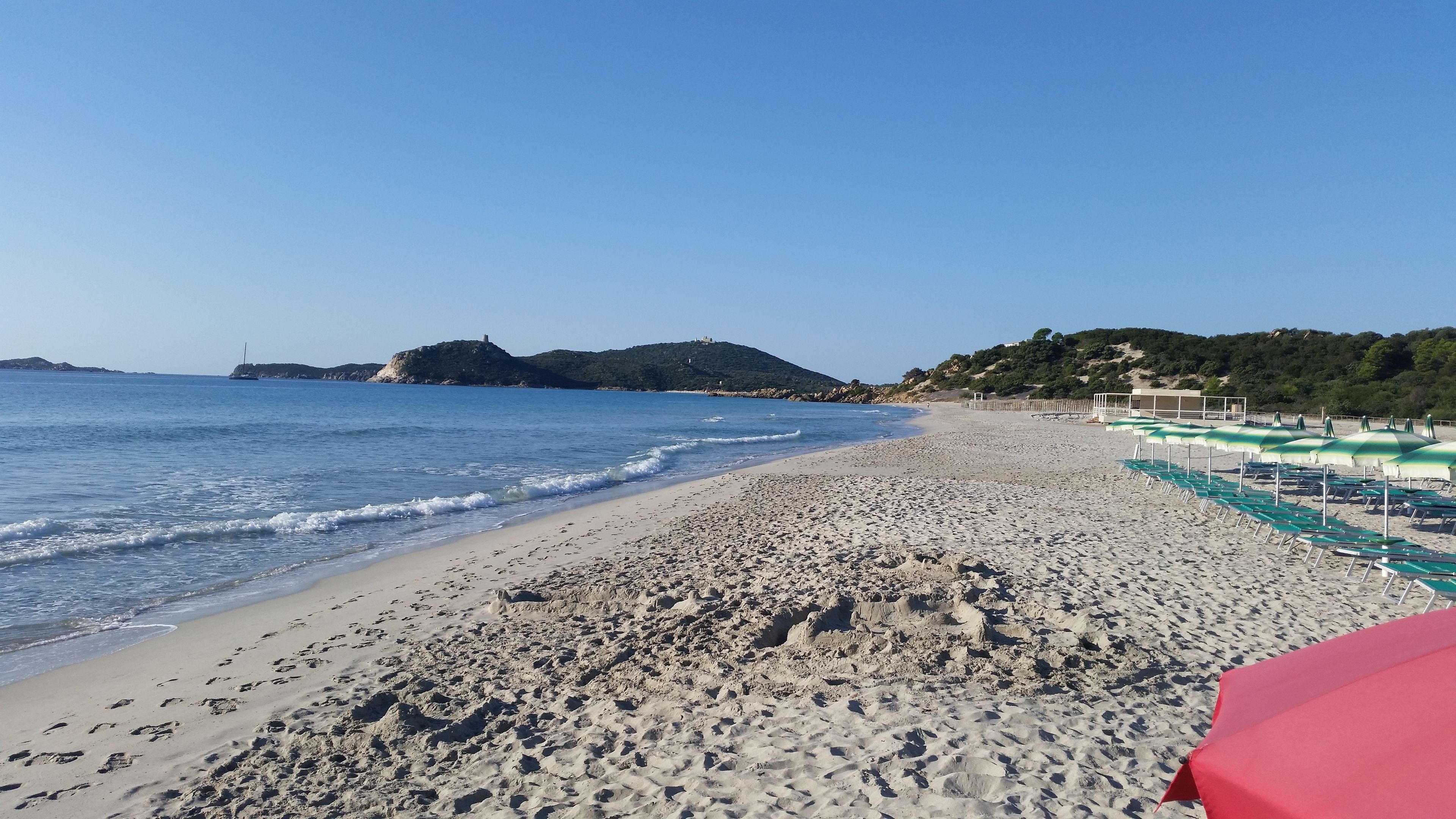 A beach with footprints, a row of green and white umbrellas, and a distant hill with a structure.