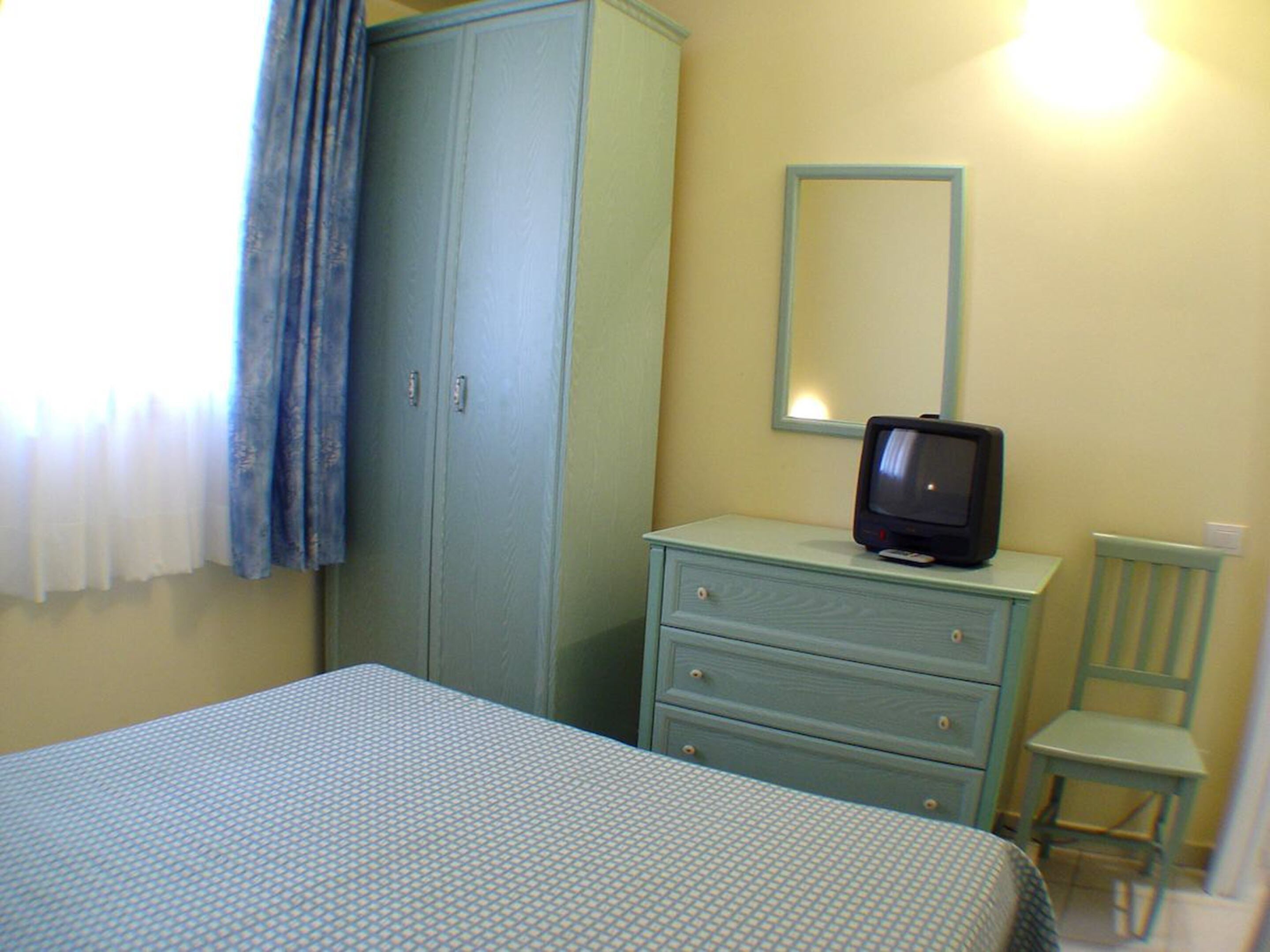 Une pièce comprenant un lit, une commode, un téléviseur posé sur le dessus et une chaise.
