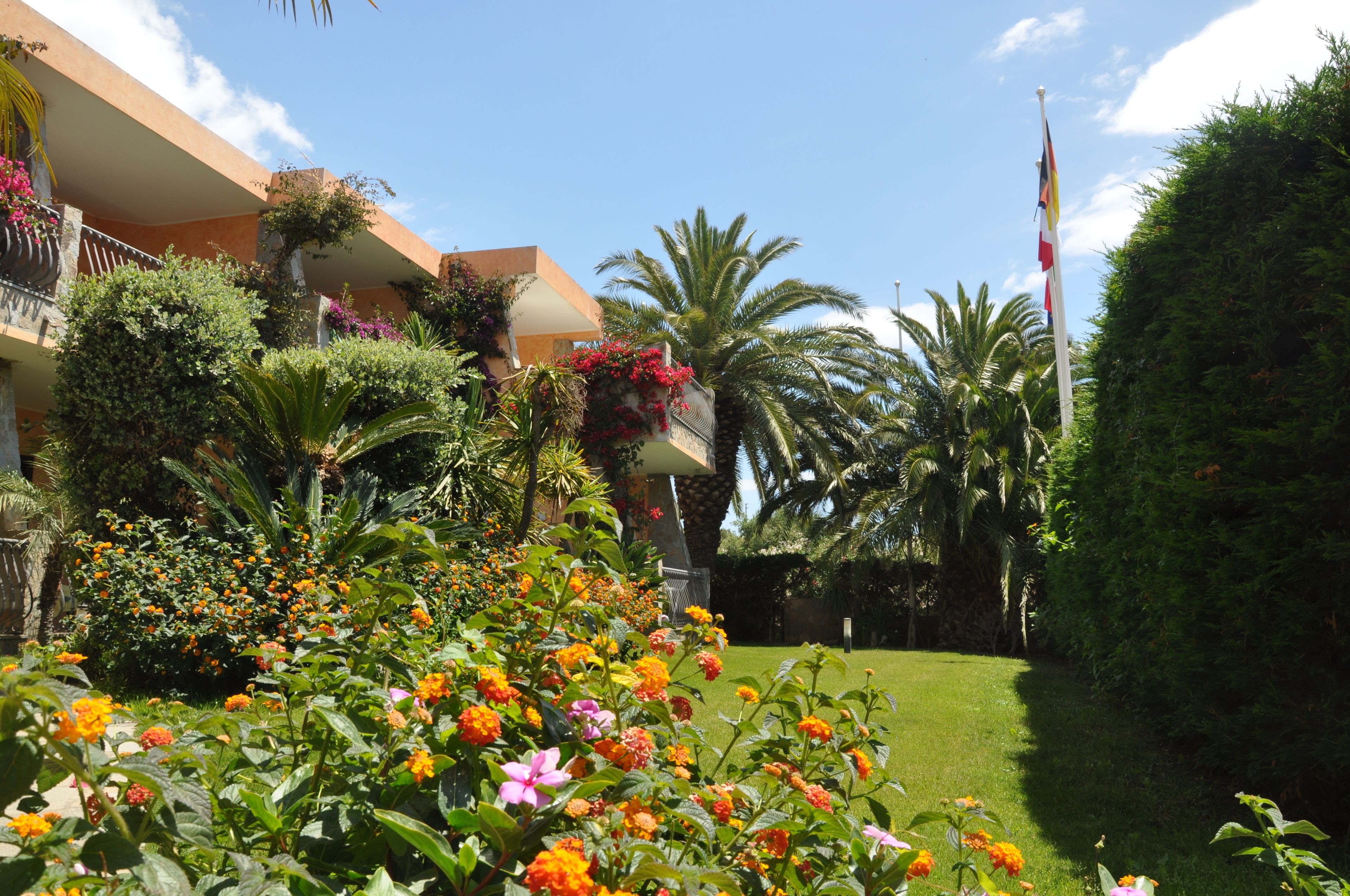 Un jardín cuidado con diversas plantas y flores, un asta de bandera y un edificio con balcones bajo un cielo despejado.