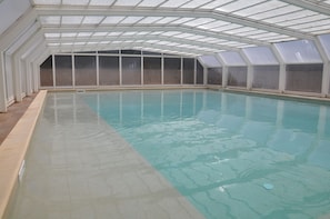 An indoor swimming pool with a glass ceiling and walls, surrounded by a tiled floor.