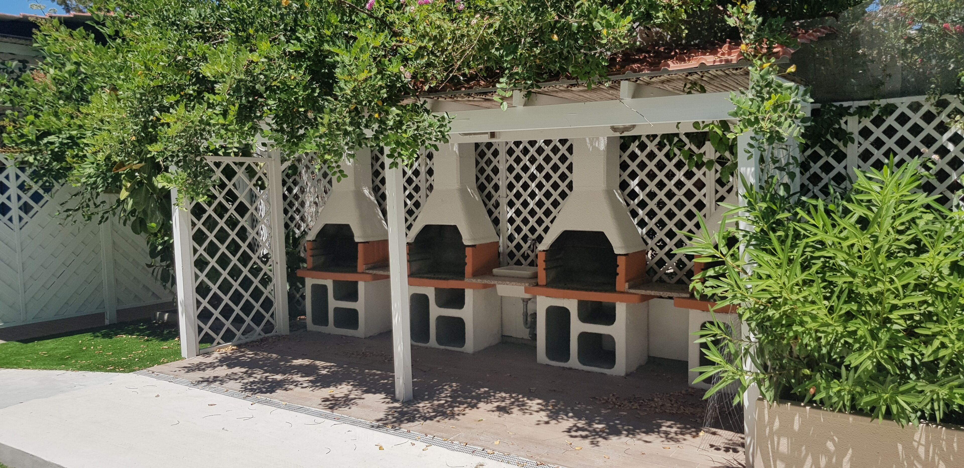 A covered outdoor seating area with benches and lattice panels, surrounded by greenery.