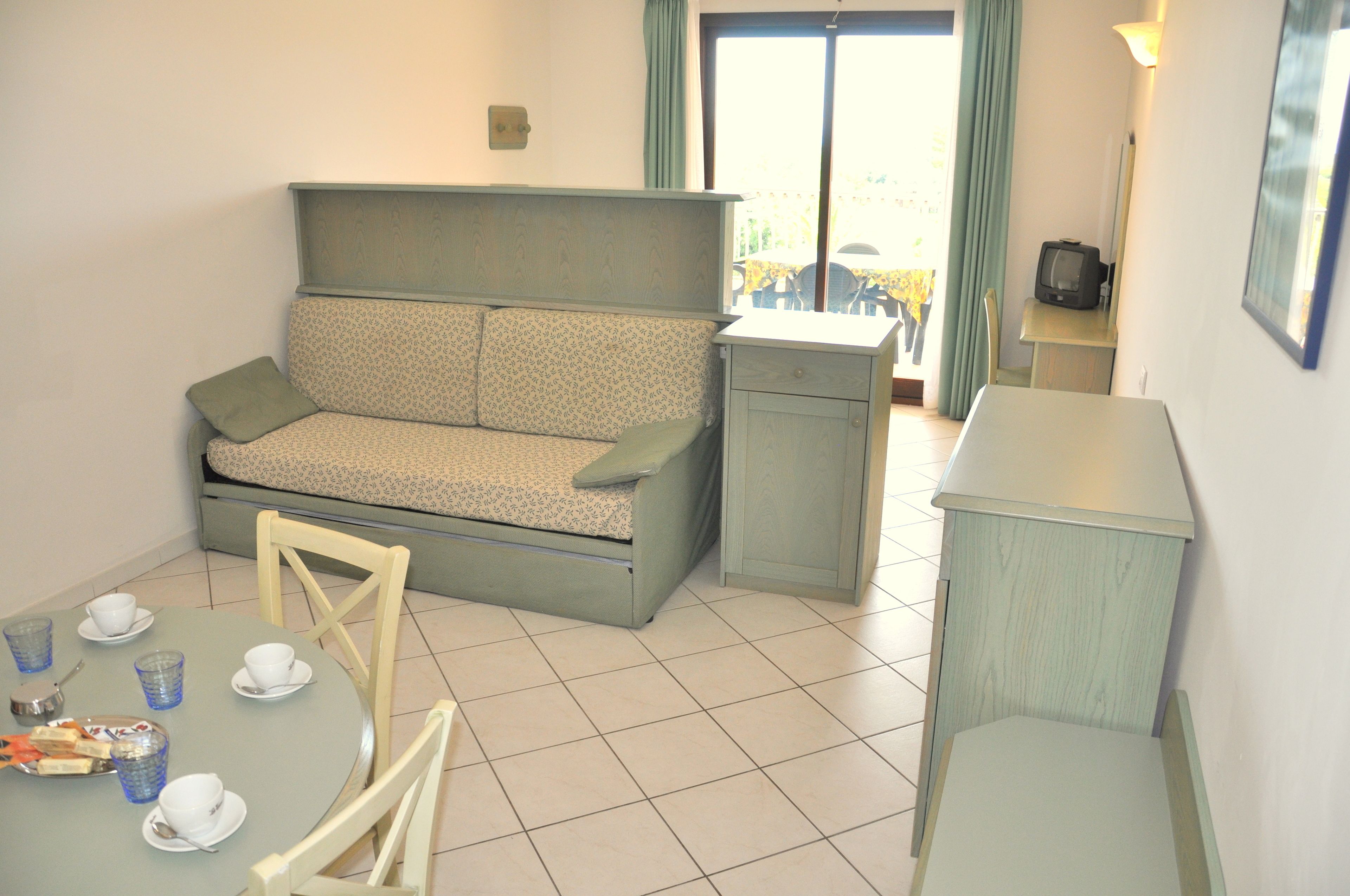 A small, compact room with a sofa, a dining table with chairs, a small cabinet, and a television.