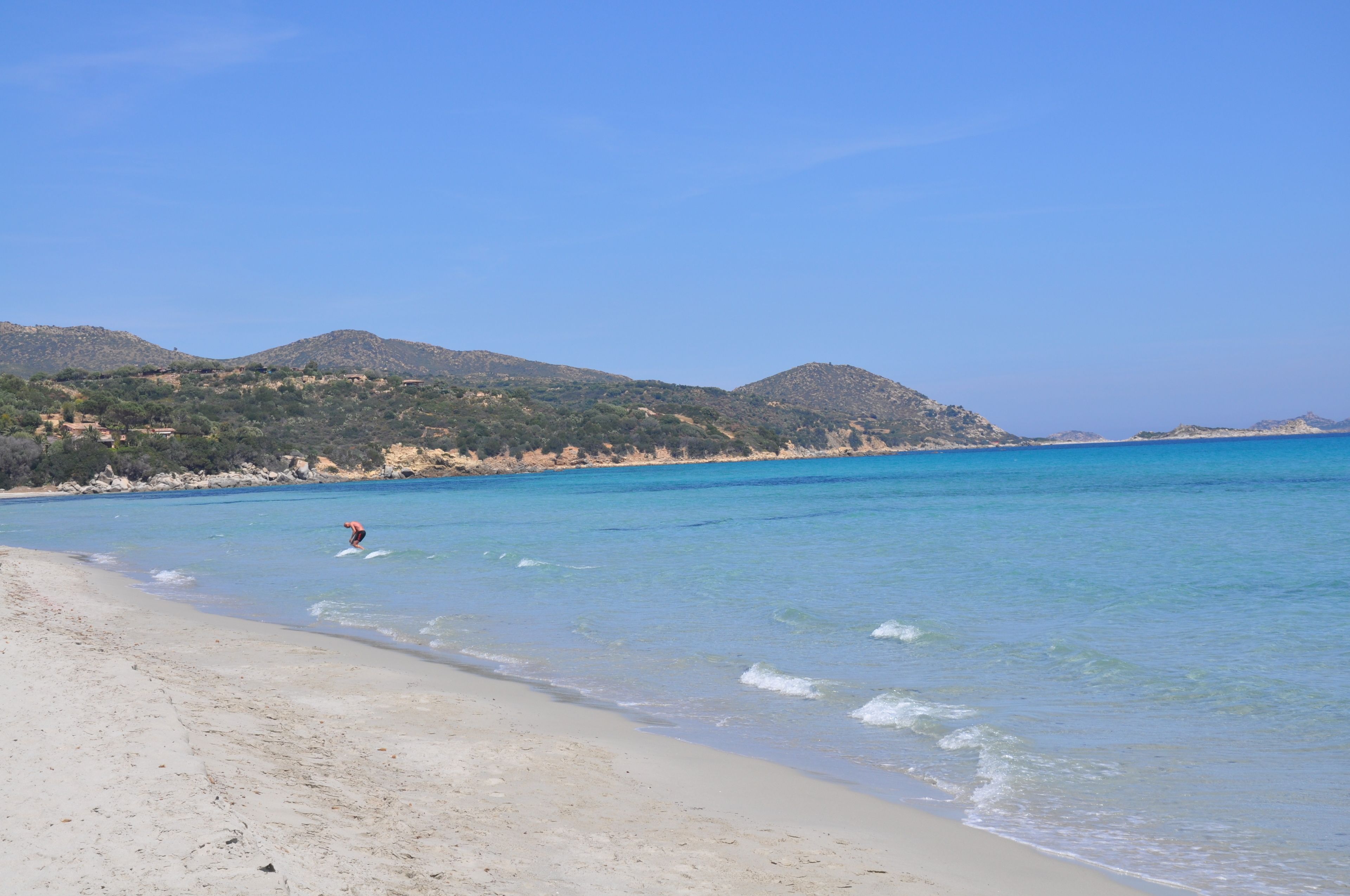 Ein Strand mit klarem, blauem Wasser, einer Person beim Surfen und Hügeln im Hintergrund.