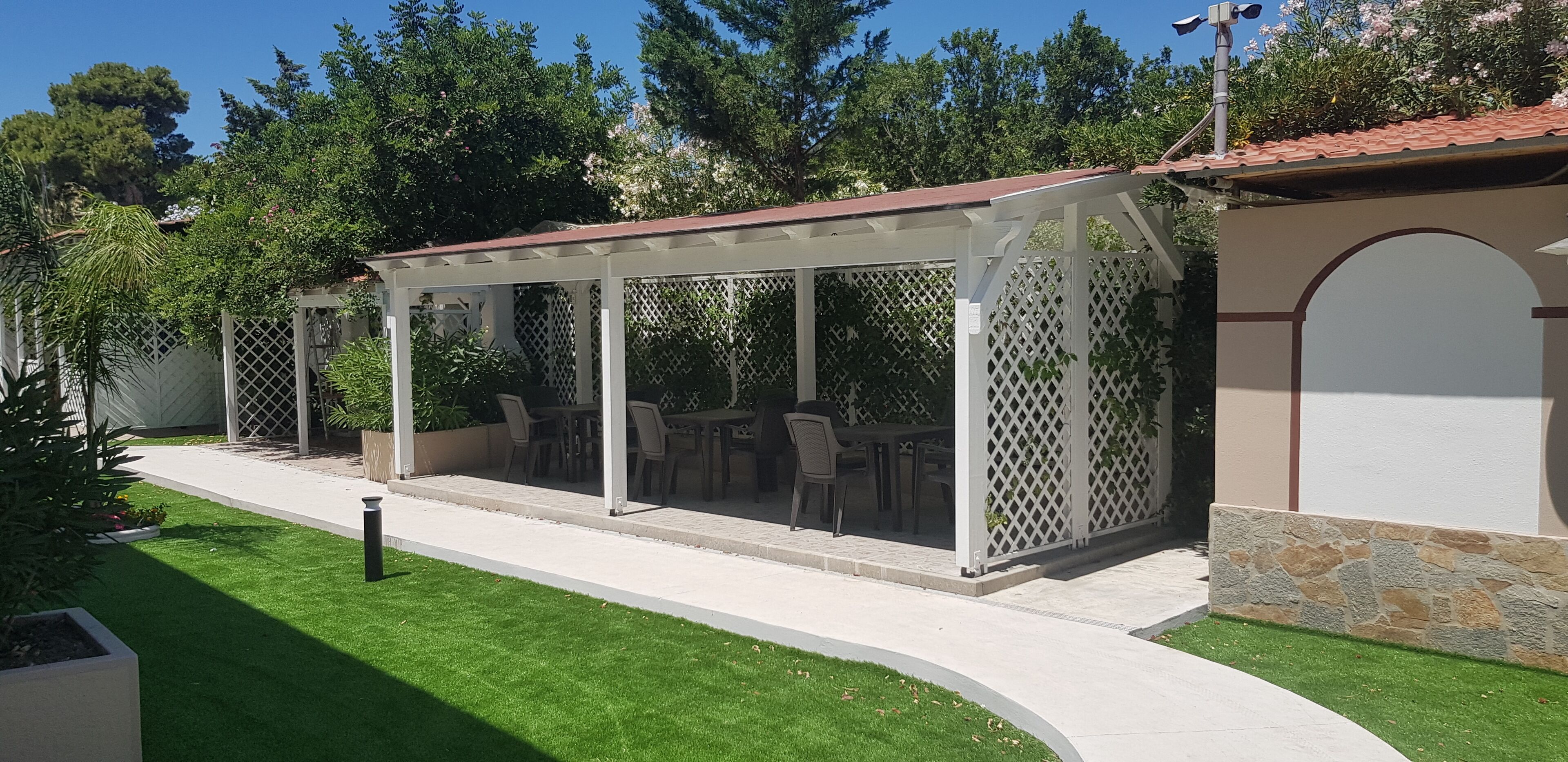 A covered outdoor seating area with tables and chairs, surrounded by a white lattice fence, set against a backdrop of trees and a building with a tiled roof.