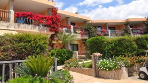 A multi-story hotel with balconies adorned with pink flowers, surrounded by lush greenery and a clear blue sky. - Palm Village (Villasimius)
