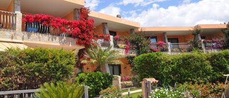 Ein mehrstöckiges Hotel mit Balkonen, die mit rosa Blumen geschmückt sind, umgeben von üppigem Grün und blauem Himmel.
