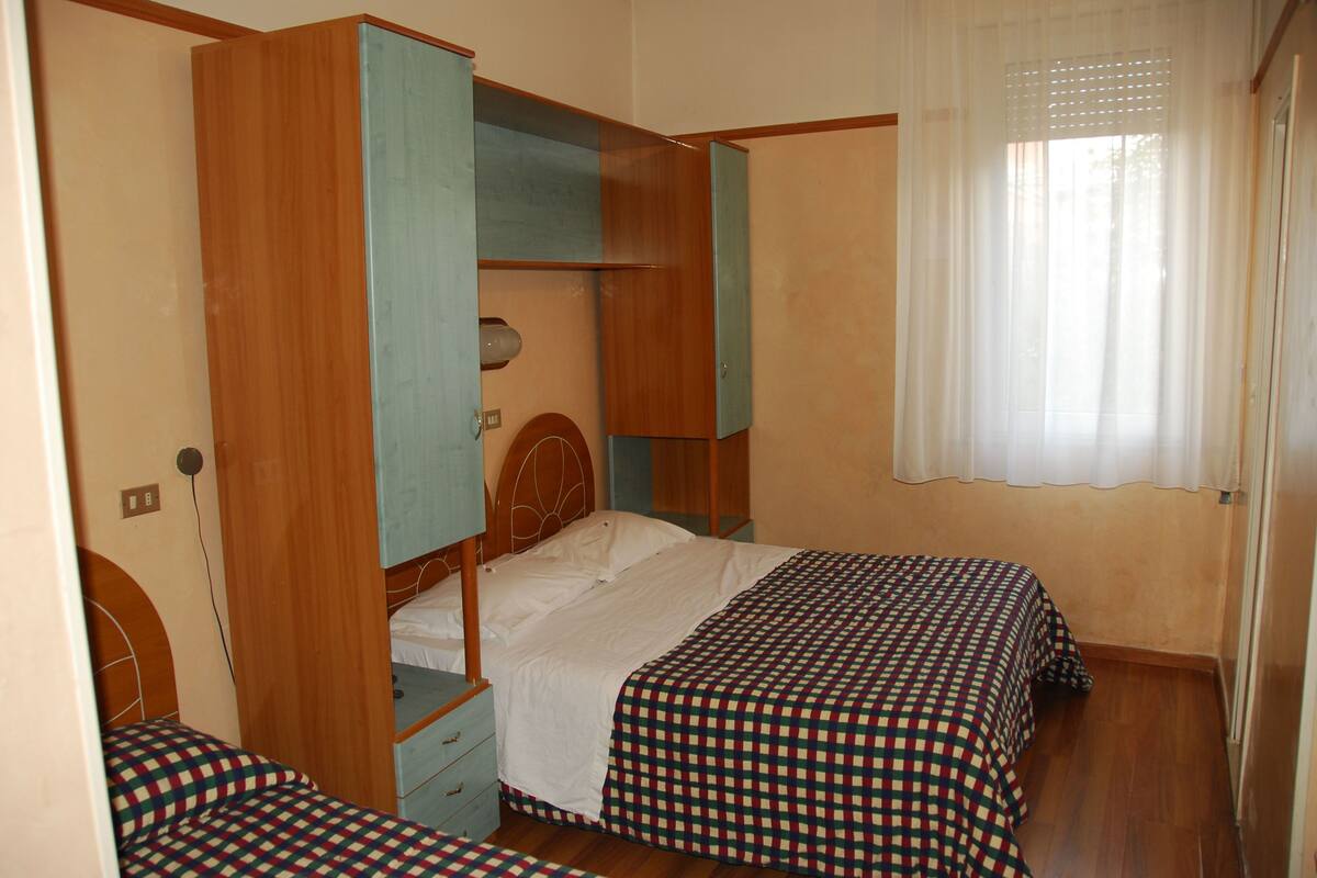 Un dormitorio con una litera, una cama individual y un ventanal con cortinas traslúcidas.