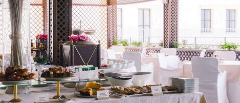 A buffet table with various pastries and desserts, including croissants and donuts, in a covered outdoor area with white chairs and tables.
