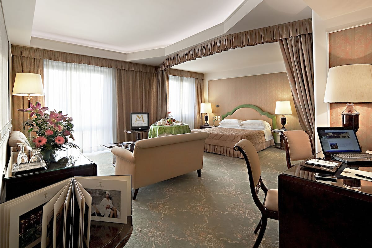 A hotel room with a bed, a desk with a laptop, a chair, a table with a lamp, and a TV.