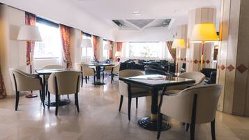 Zona pranzo di un hotel con diversi tavoli e sedie, ampie finestre con tende e lampade da terra.