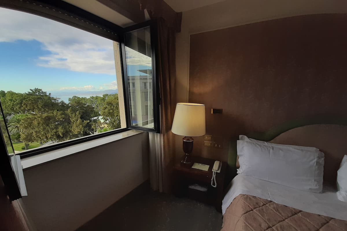 A hotel room with a bed, a nightstand, a lamp, a window with a view of trees and a building, and a telephone.