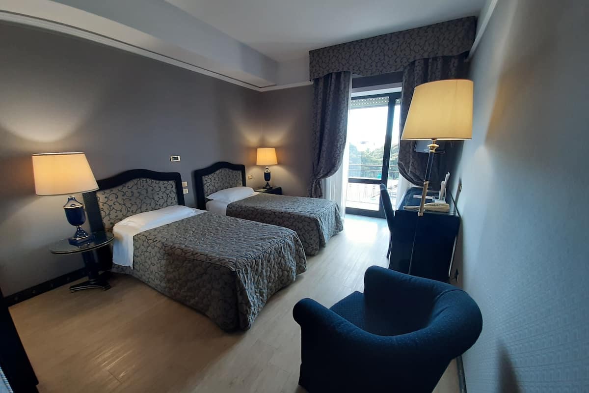 A hotel room with two beds, a chair, and a balcony with curtains.