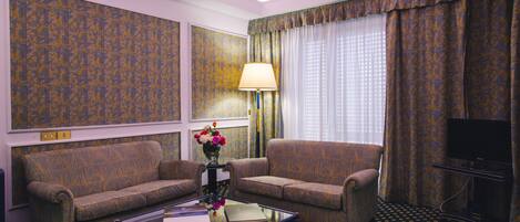 Habitación de hotel con dos sofás estampados, una mesa de centro de cristal con un libro y flores, una lámpara, un televisor y cortinas con diseño.