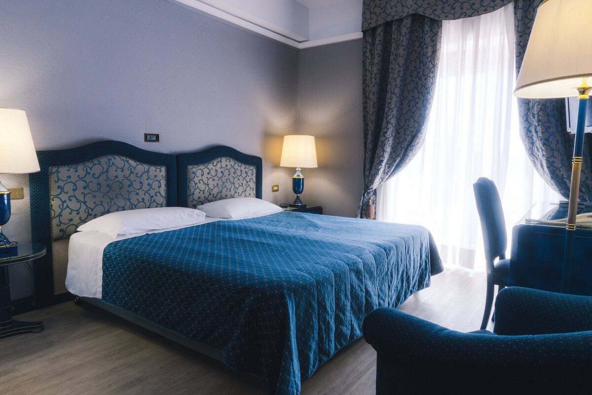 A hotel room with a large bed, two bedside tables with lamps, a chair, and a window with curtains.
