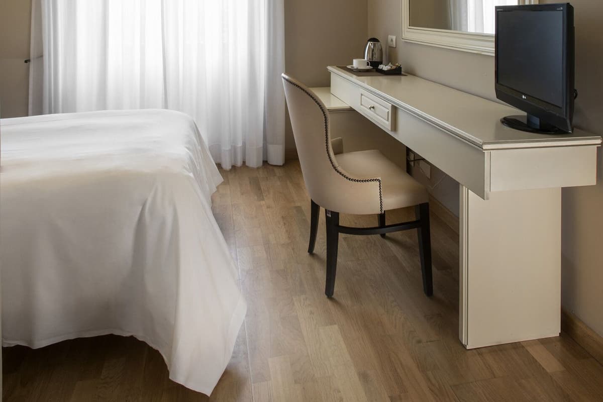 Habitación de hotel con cama, escritorio, silla, televisión y un ventanal con cortinas.